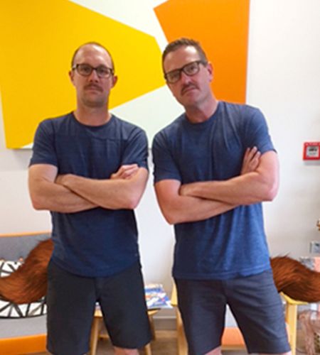 Two men standing wearing glasses and same dark blue outfits