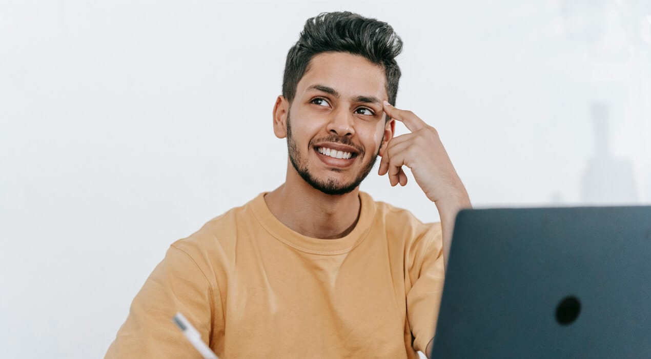 Smiling young man sitting at a desk, with a laptop in front of him and pen in hand